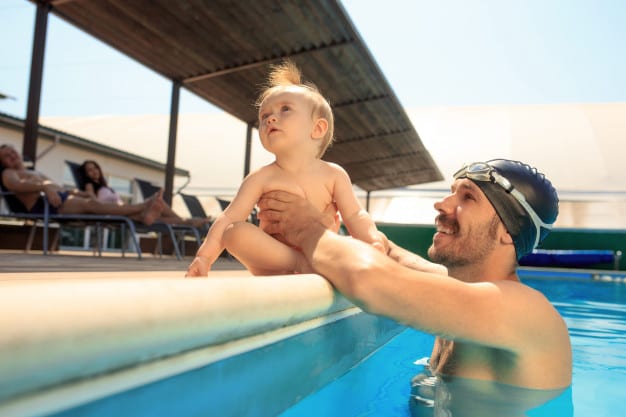 baby sitting by the poolside as guided by father